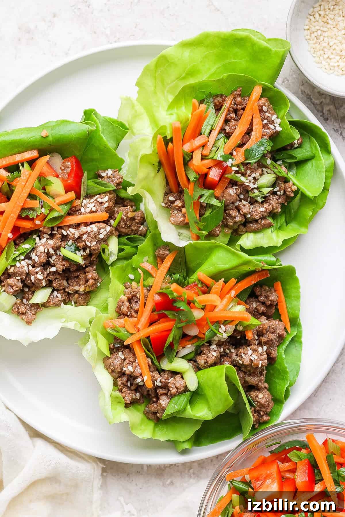 Korean Beef Lettuce Wraps with Gochujang Glaze 2 Four lettuce wraps filled with ground meat, shredded carrots, diced red bell peppers, and green onions, served on a white plate.