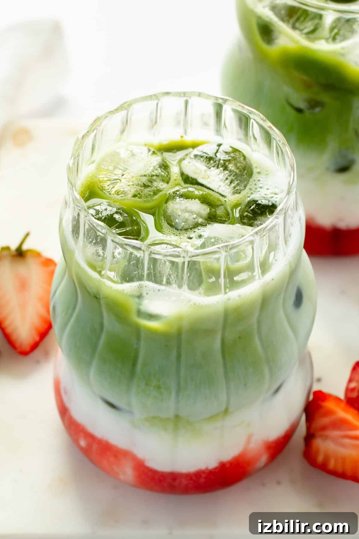 Iced Strawberry Matcha Protein Smoothie Recipe 2 Layered matcha latte with ice, featuring green matcha, white milk, and red strawberry syrup. Served in a clear, textured glass with sliced strawberries on the side.