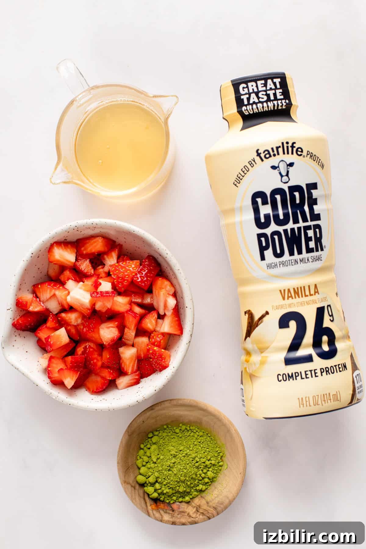 Iced Strawberry Matcha Protein Smoothie Recipe 3 Vanilla protein milkshake bottle next to a small jug of liquid, a bowl of chopped strawberries, and a bowl of matcha powder on a white surface.