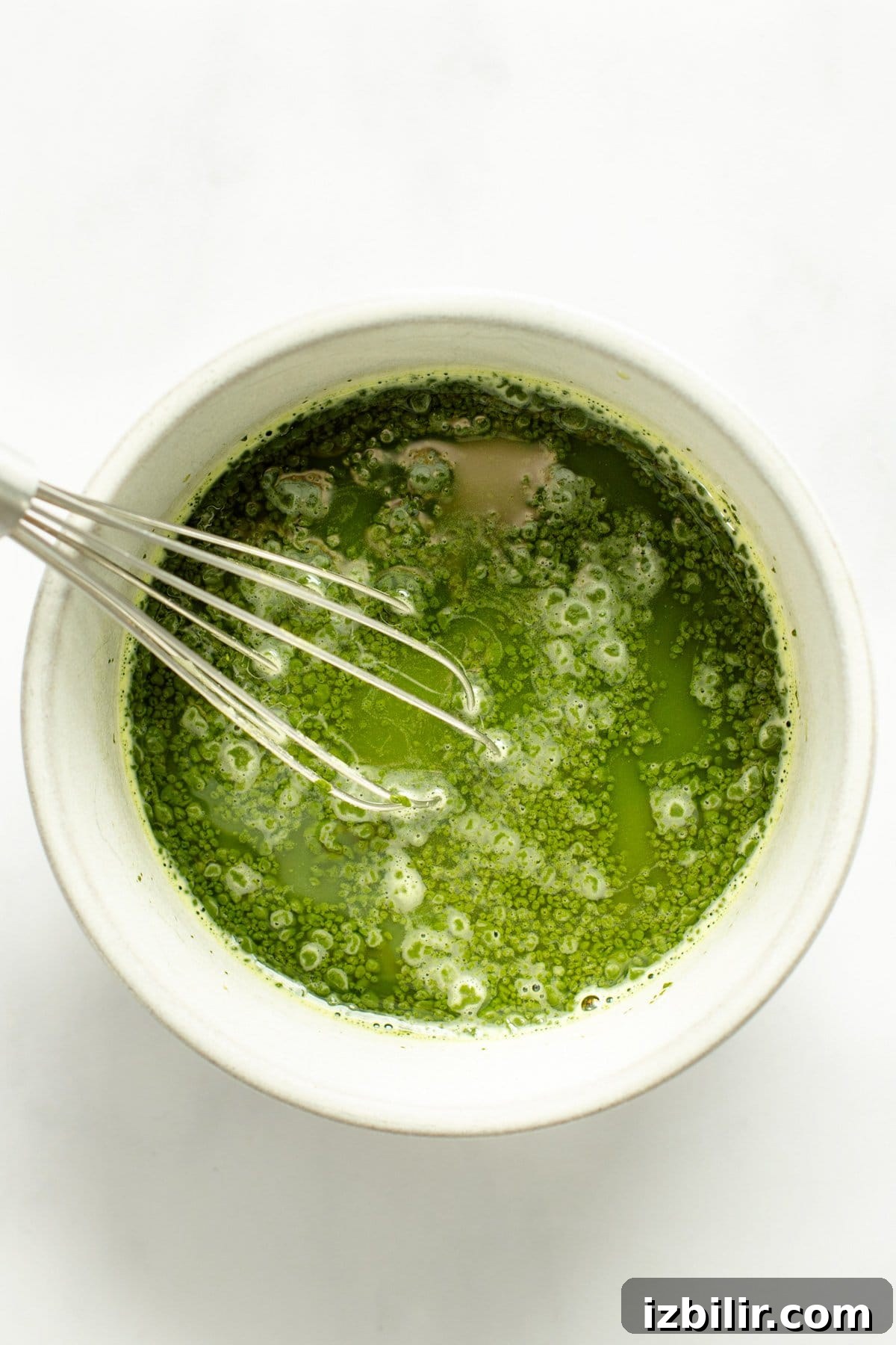 Iced Strawberry Matcha Protein Smoothie Recipe 4 A whisk in a white bowl containing frothy green matcha tea against a white background.