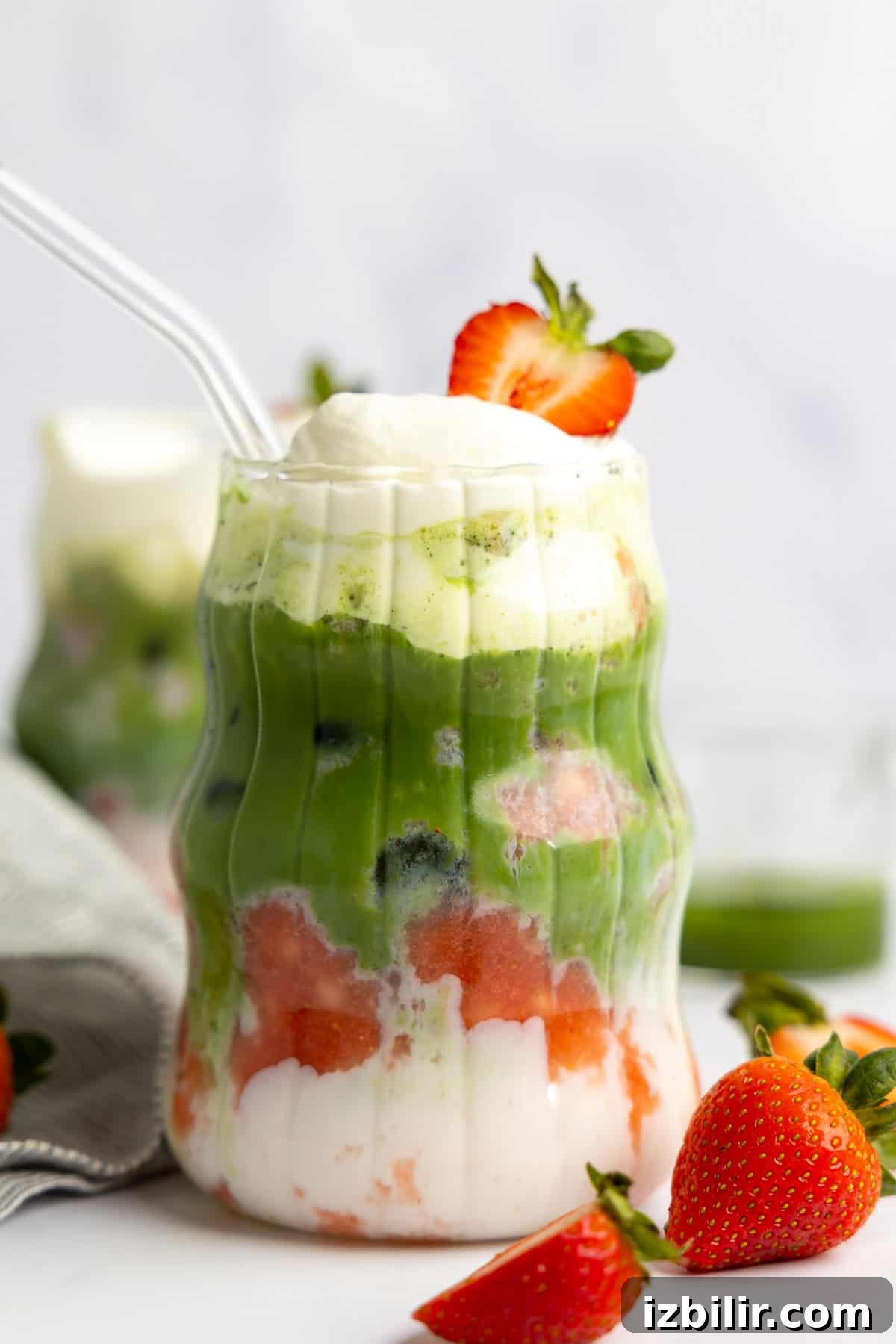 Iced Strawberry Matcha Protein Smoothie Recipe 6 A layered drink featuring protein milk, green matcha, vibrant fruit pieces, and whipped cream in a clear glass. It's elegantly topped with a strawberry slice and accompanied by a transparent straw.