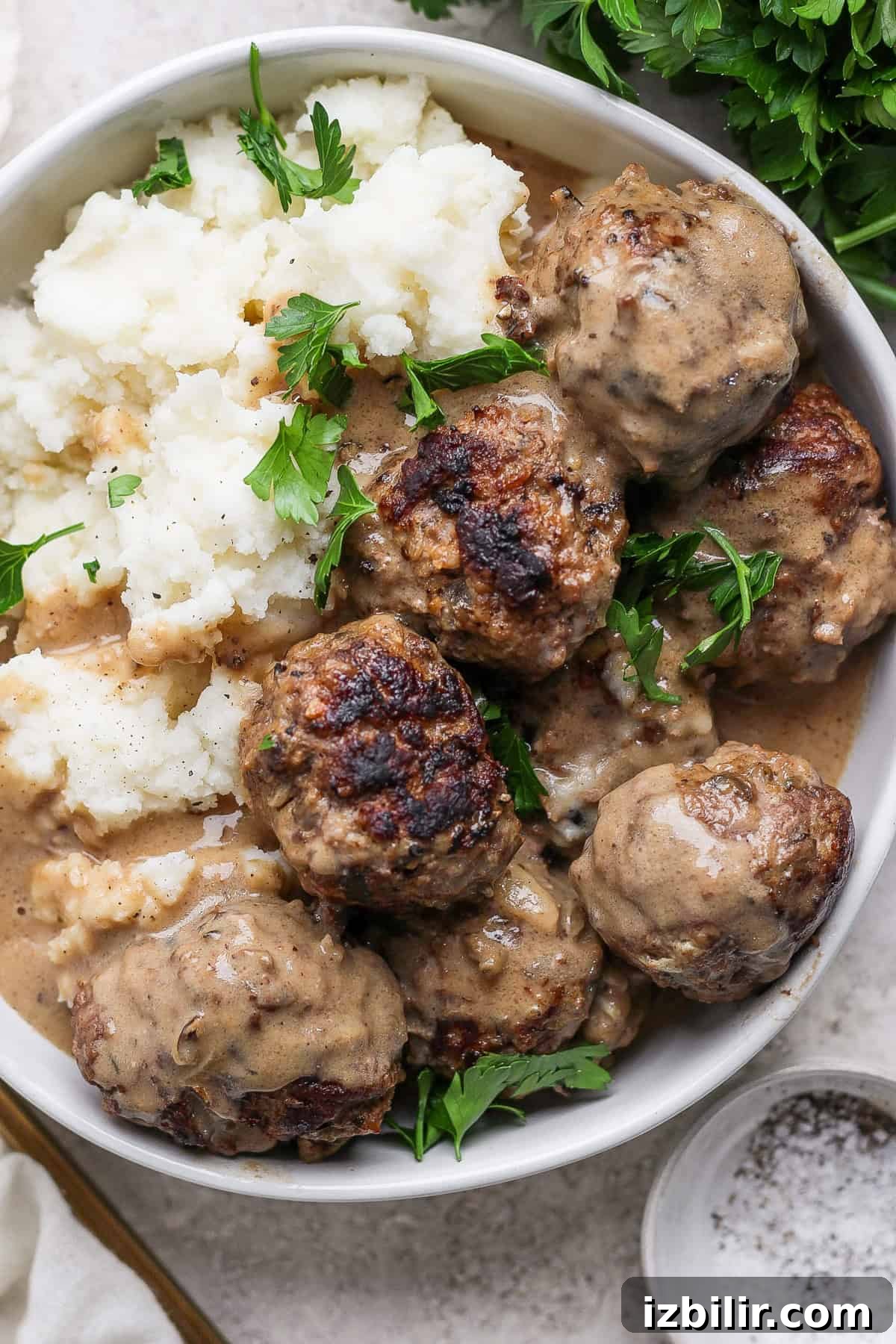 Minnesota-Style Swedish Meatballs with Creamy Gravy 2 A bowl of meatballs in creamy sauce next to a serving of mashed potatoes. Fresh parsley garnishes the dish.