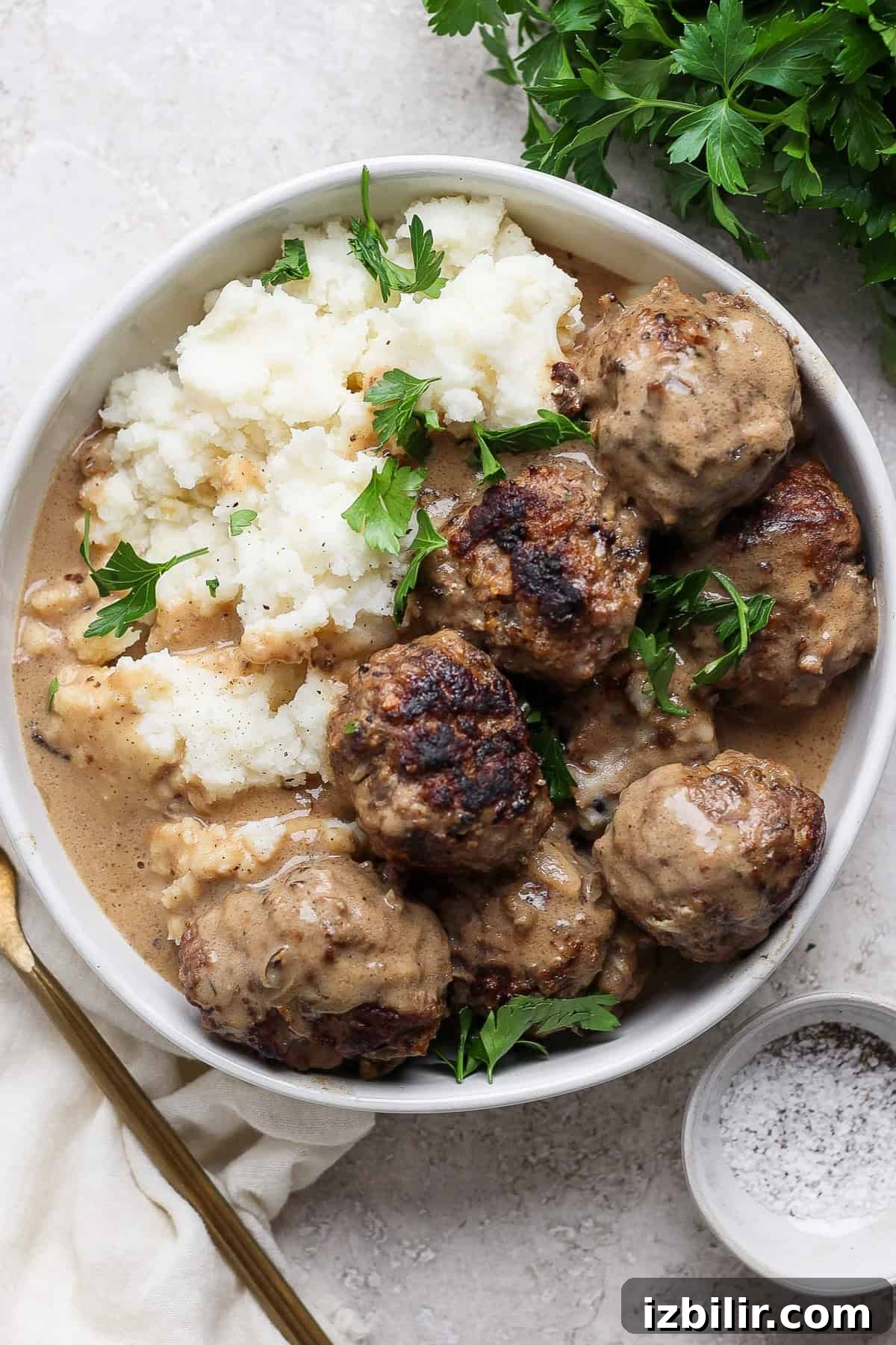 Minnesota-Style Swedish Meatballs with Creamy Gravy 5 A plate of meatballs covered in gravy, served with mashed potatoes and garnished with fresh parsley. A fork and a small bowl of pepper sit adjacent to the plate.