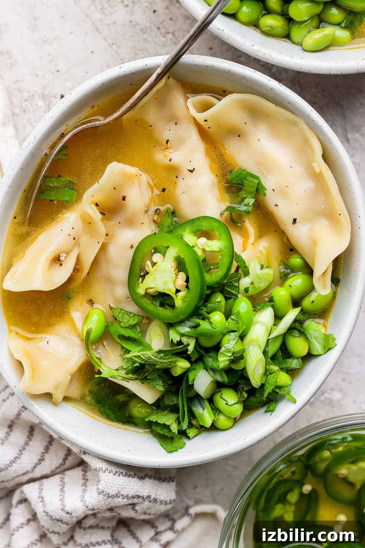 Zesty Lemon Wonton Soup Recipe 2 A bowl of broth with dumplings, sliced green chili, edamame, and chopped green onions, garnished with herbs. A spoon is resting in the bowl.