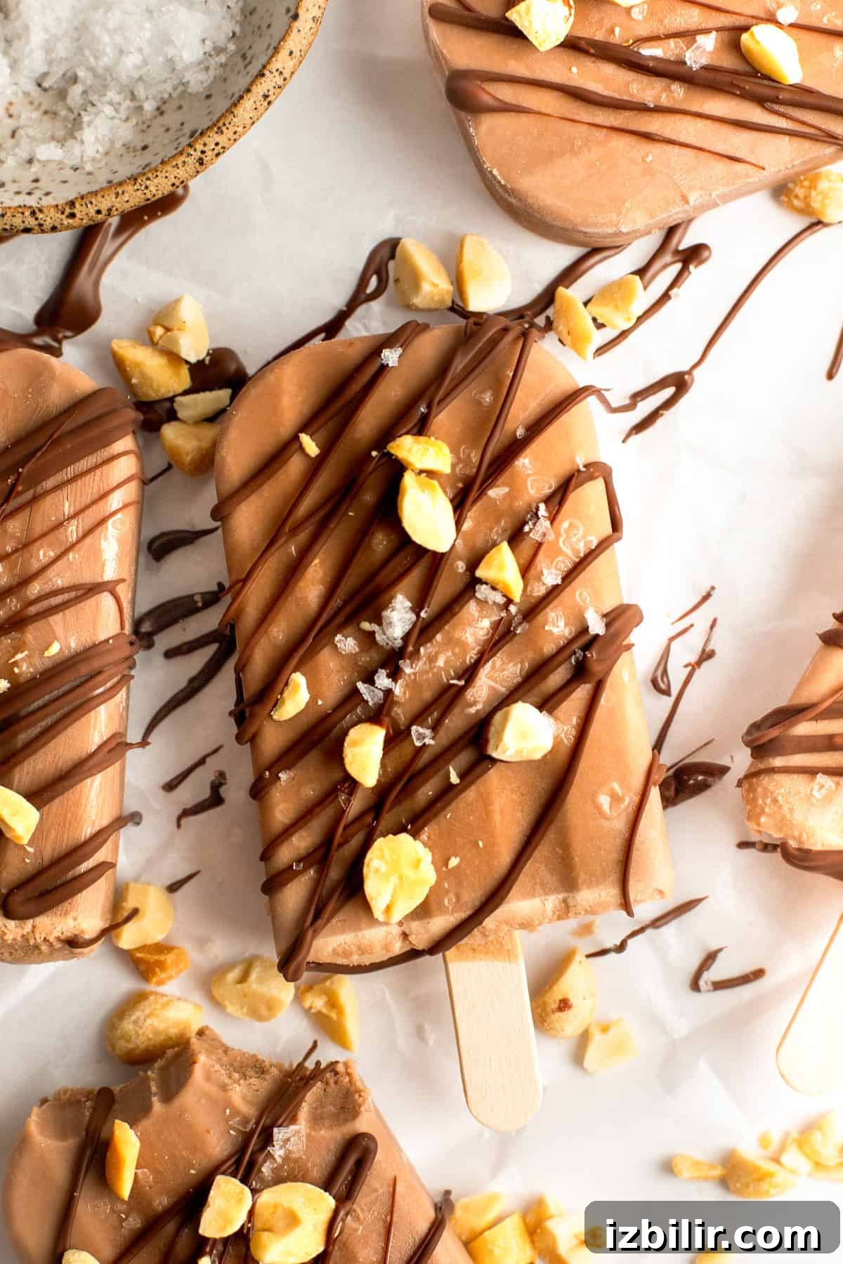 Chocolate popsicles drizzled with chocolate and sprinkled with chopped nuts and sea salt on a white surface.