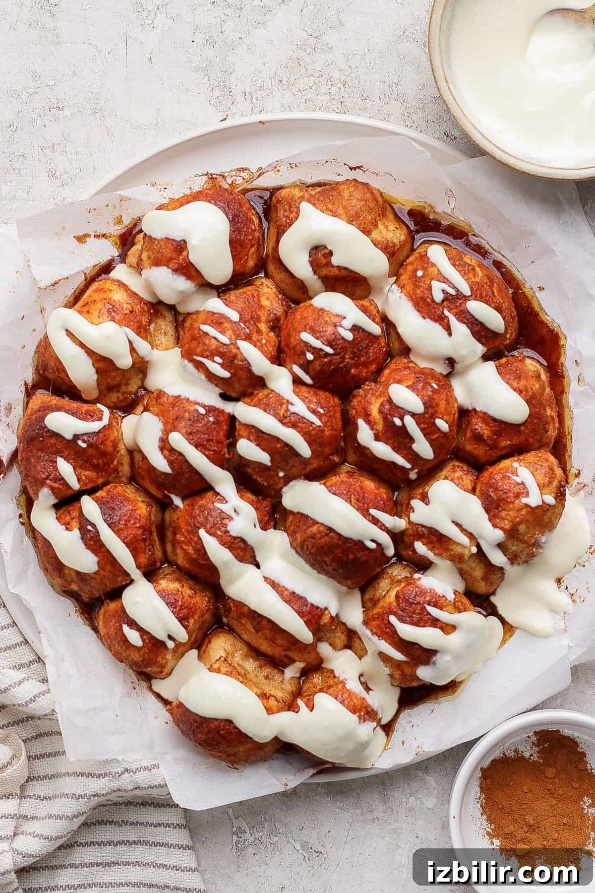 Air Fryer Protein Monkey Bread Recipe 2 A round cinnamon pull-apart bread topped with cream cheese icing on parchment paper. Nearby are a bowl of icing and a dish of cinnamon.