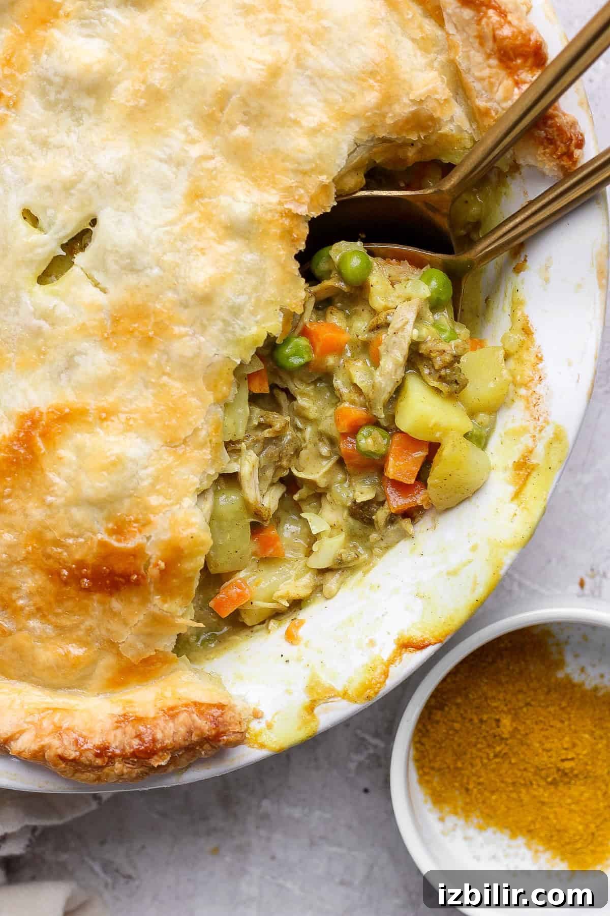 Curried Chicken Pot Pie Recipe with Flaky Crust 2 A pot pie with a flaky crust partially open, revealing a filling of chicken, carrots, peas, and potatoes. Two spoons are placed inside, next to a small dish of curry powder.