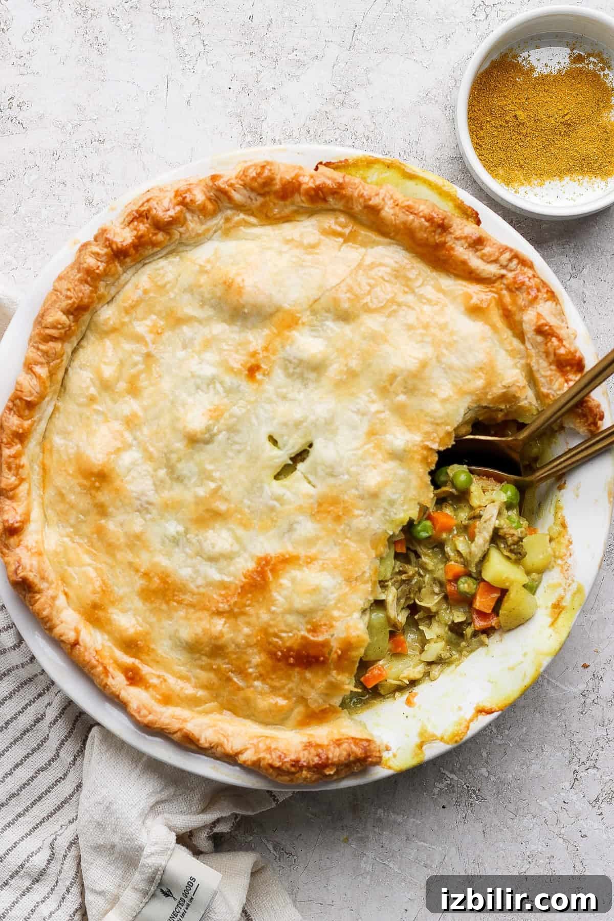 Curried Chicken Pot Pie Recipe with Flaky Crust 6 A savory pie with a golden crust is partially sliced, revealing a filling of vegetables and chicken. Two spoons are in the pie, and a bowl of seasoning is nearby.