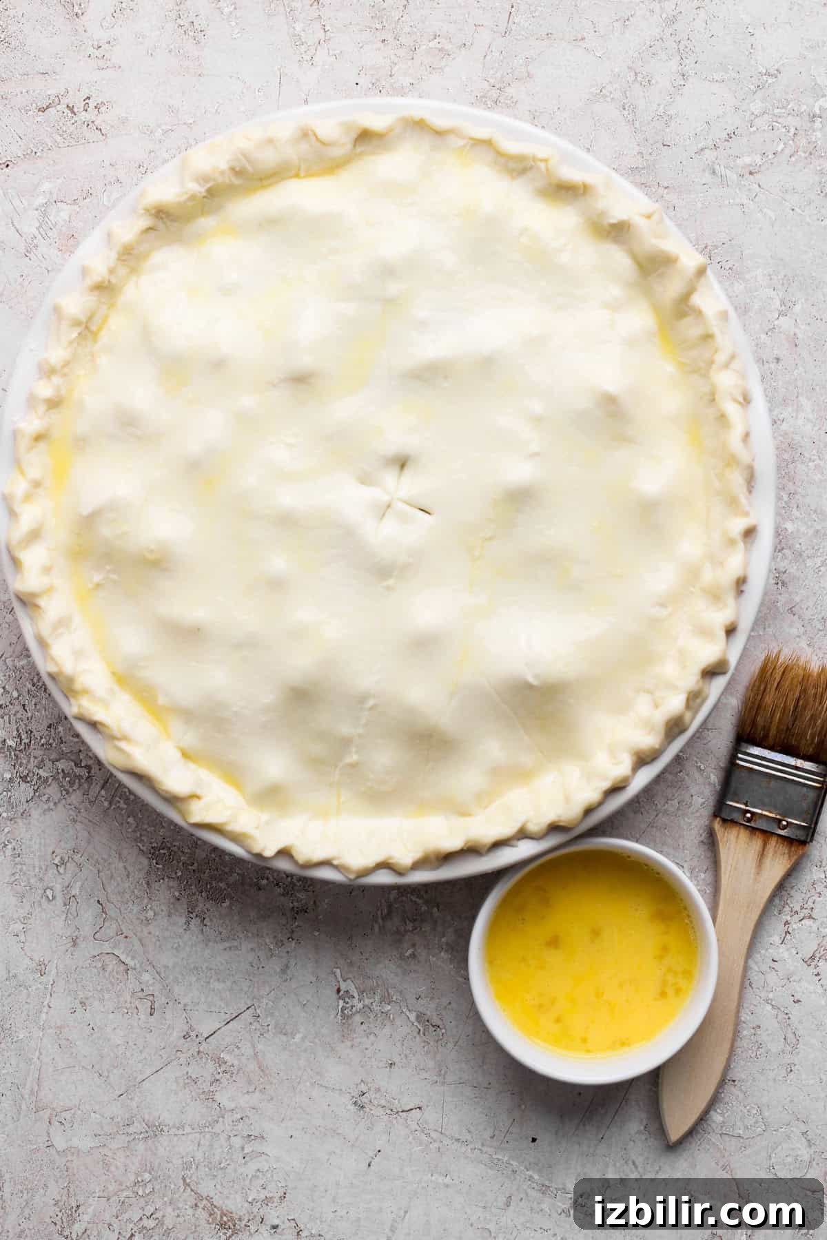 Curried Chicken Pot Pie Recipe with Flaky Crust 3 Unbaked pie with a crimped edge on a round dish, accompanied by a small bowl of egg wash and a pastry brush on a textured surface.