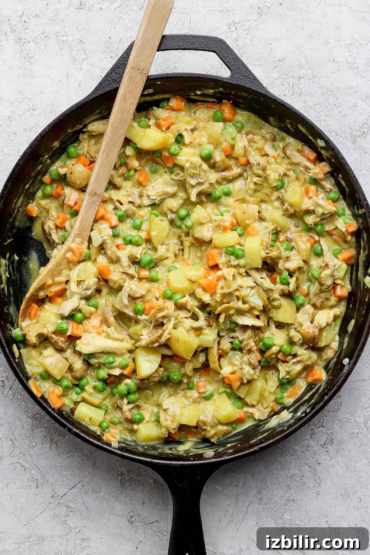 Curried Chicken Pot Pie Recipe with Flaky Crust 4 A skillet of cooked chicken, potatoes, carrots, and peas mixed in a creamy sauce, with a wooden spoon on the side.