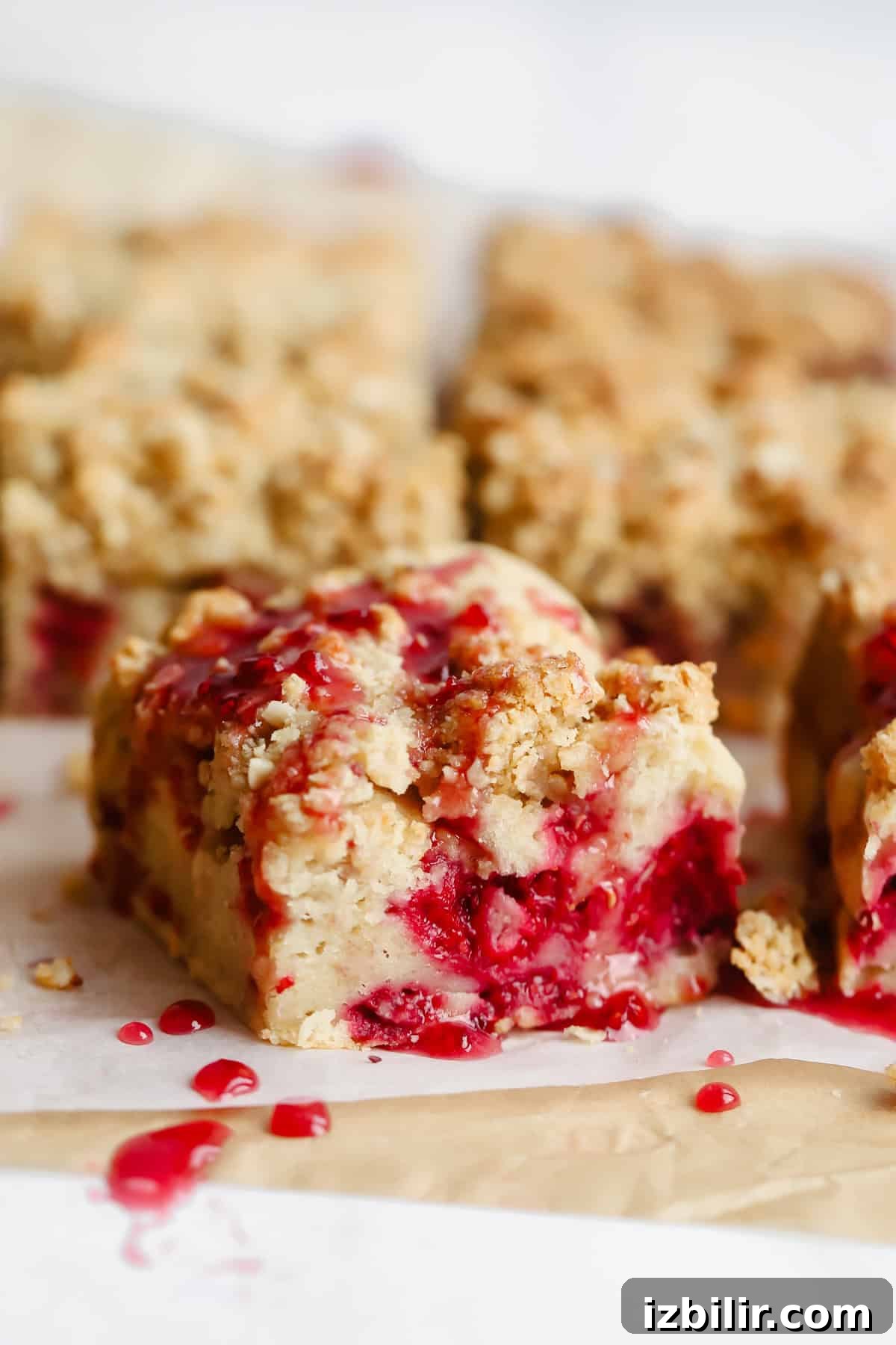 Close-up of crumbly dessert bars with a raspberry filling, topped with a drizzle of red sauce, arranged on parchment paper.