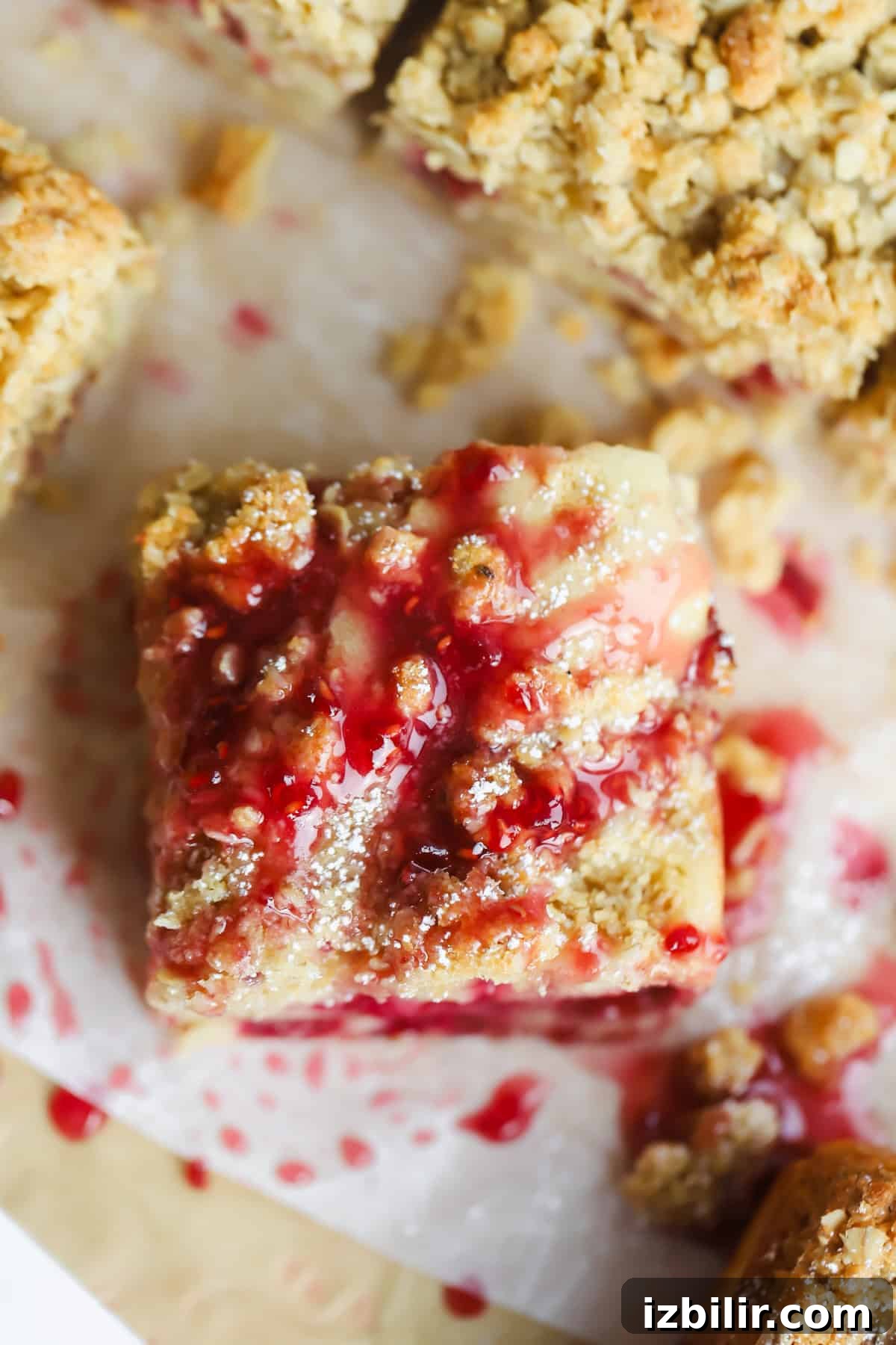 A square piece of dessert topped with a crumbly streusel and drizzled with red fruit sauce on parchment paper.