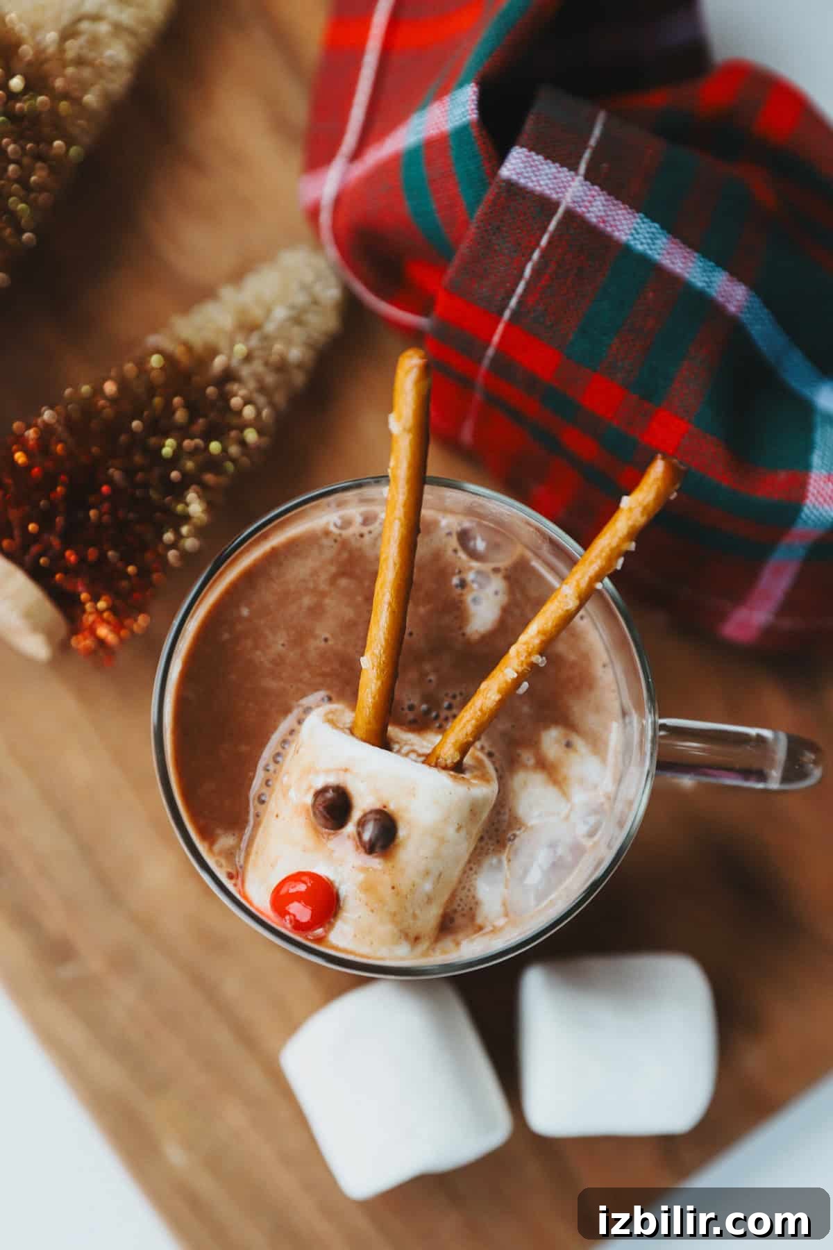 Melting Snowman Whipped Cream Bombs Recipe 2 A cup of hot chocolate with a marshmallow decorated as a reindeer using pretzels, candy eyes, and a red candy nose, next to a plaid cloth and mini Christmas trees.