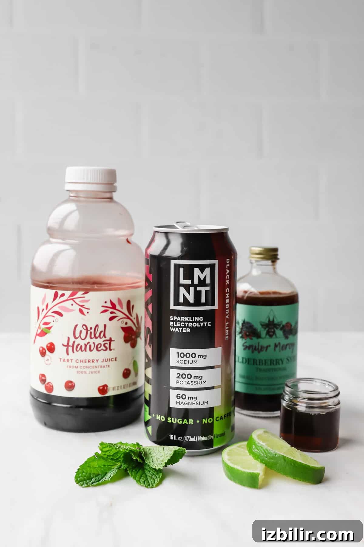 Sleepy Girl Mocktail Recipe: Soothing Caffeine-Free Nightcap 3 A bottle of tart cherry juice, a can of LMNT electrolyte drink, elderberry syrup, mint leaves, and lime slices on a white surface.