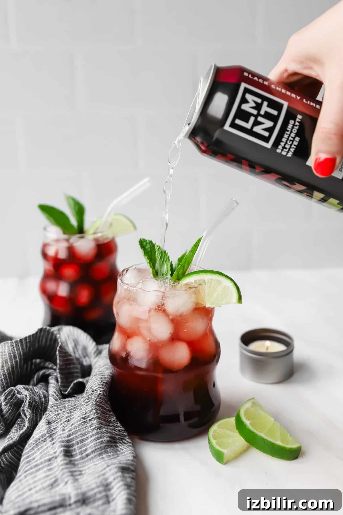 Sleepy Girl Mocktail Recipe: Soothing Caffeine-Free Nightcap 5 A hand pours a black cherry drink into a glass filled with ice, cherries, lime slices, and mint. Another similar drink is in the background, next to lime wedges and a small candle.