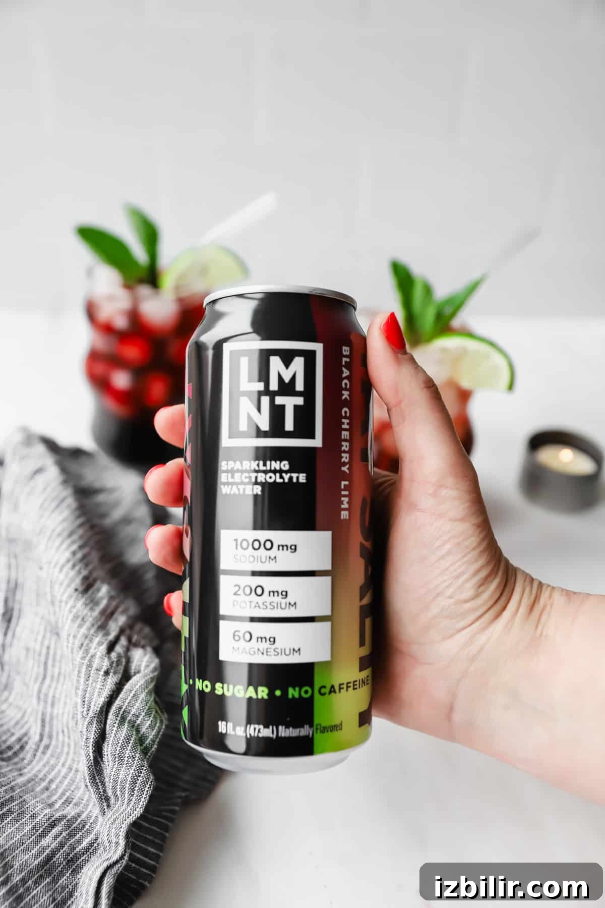 Sleepy Girl Mocktail Recipe: Soothing Caffeine-Free Nightcap 6 Hand holding a can of LMNT black cherry lime sparkling electrolyte water. Two glasses with red drinks and lime wedges are in the background.