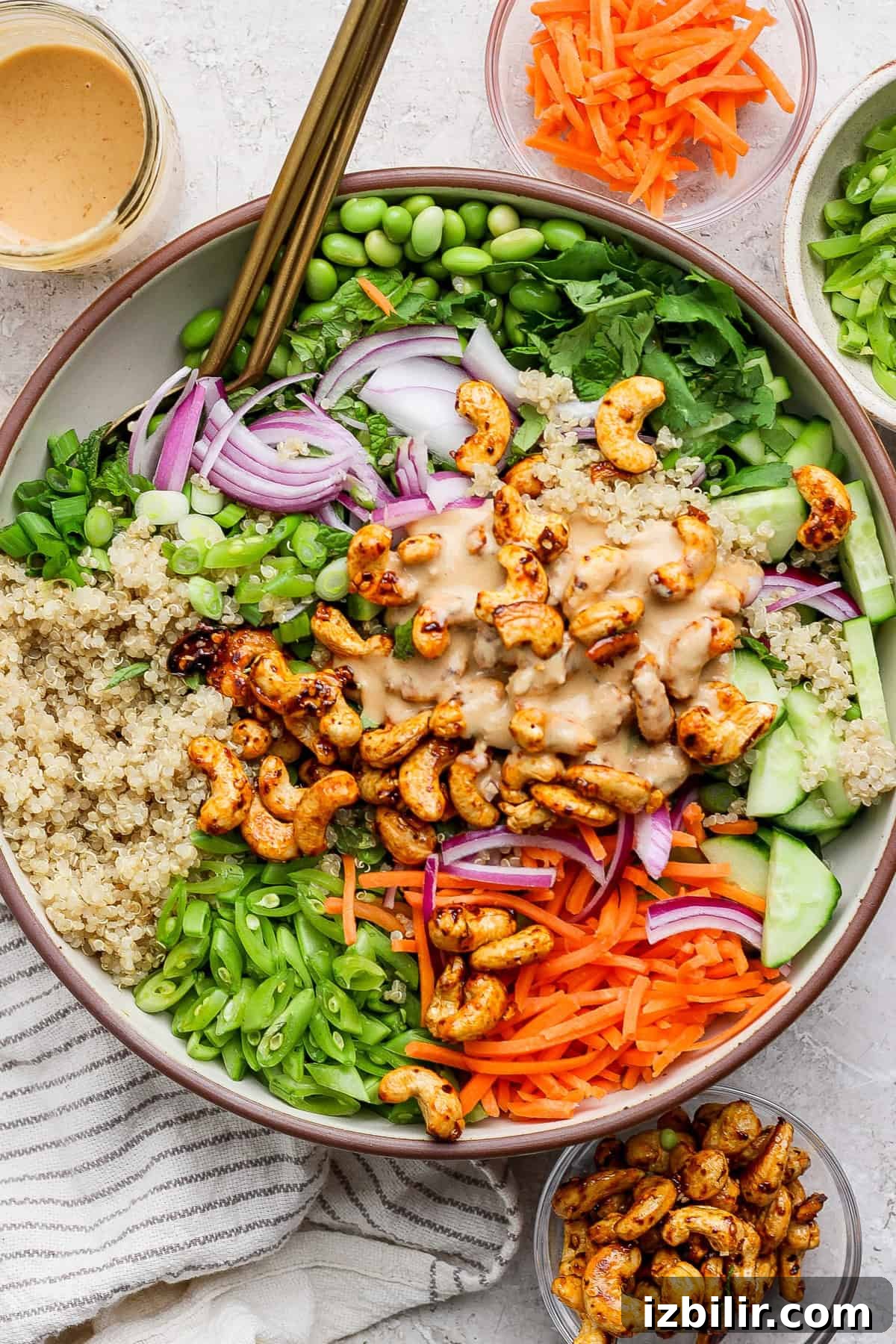 Crunchy Cashew and Edamame Salad with Sesame Dressing 2 A bowl containing quinoa, cashews, sliced red onions, carrots, cucumbers, edamame, and greens, topped with a creamy dressing. Additional ingredients are in small bowls around it.