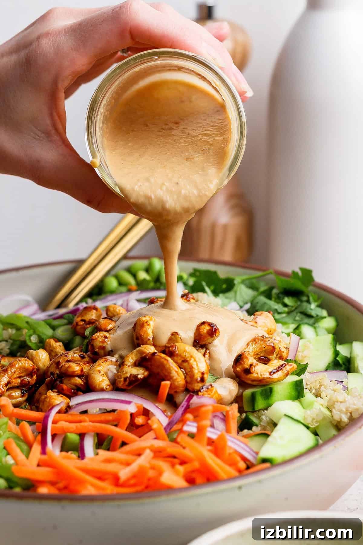 Crunchy Cashew and Edamame Salad with Sesame Dressing 4 A hand pours creamy dressing over a salad with carrots, cucumbers, red onions, roasted chickpeas, and greens in a bowl.