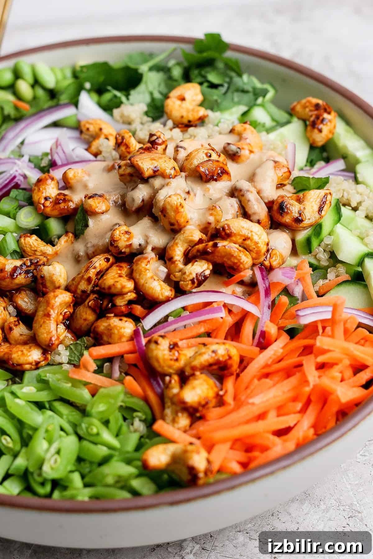 Crunchy Cashew and Edamame Salad with Sesame Dressing 5 A colorful salad with cashews, quinoa, cucumbers, red onions, sliced carrots, green beans, and greens, topped with a creamy dressing, served in a bowl.