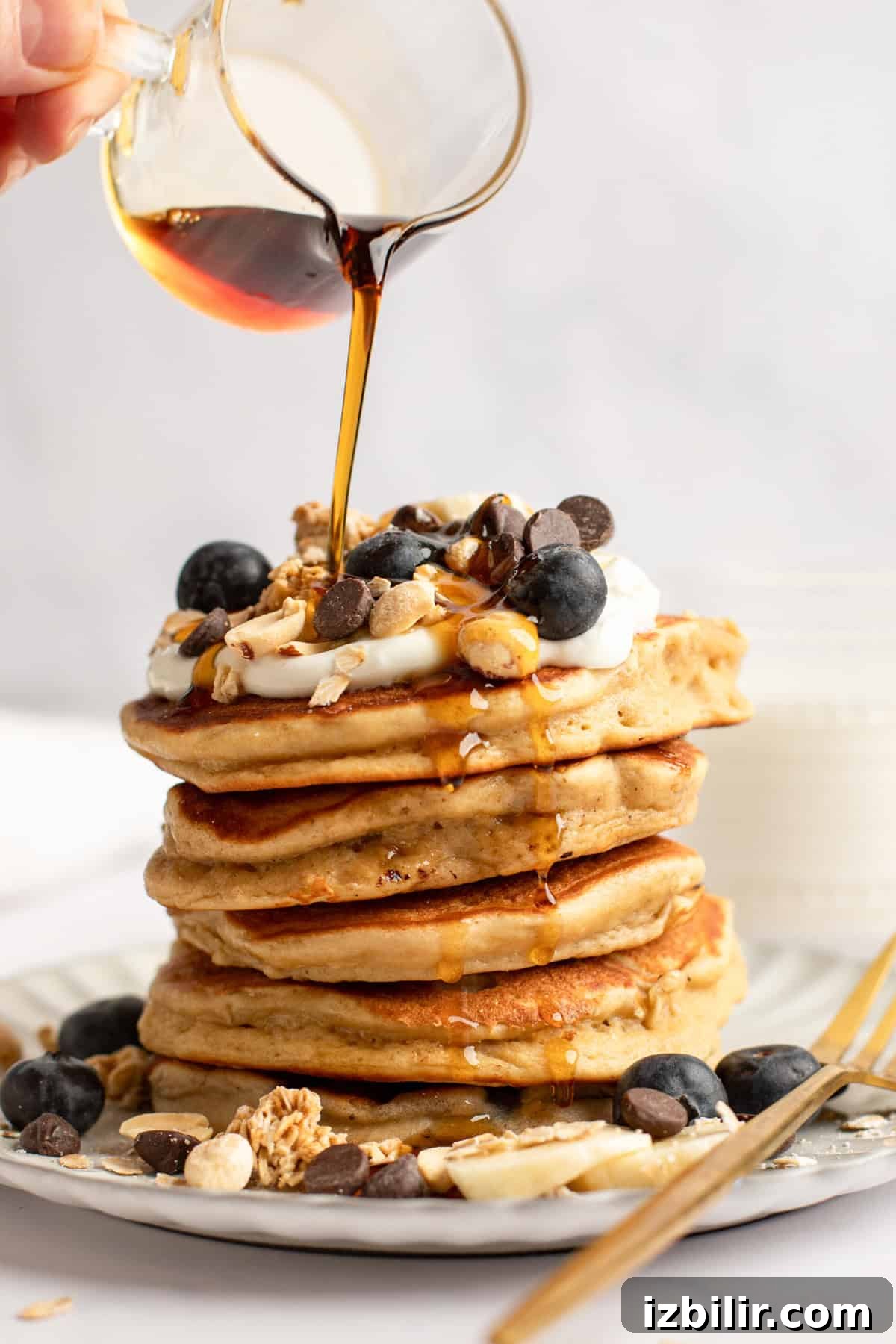 A stack of pancakes topped with blueberries, chocolate chips, granola, and nuts. Syrup is being poured over them.
