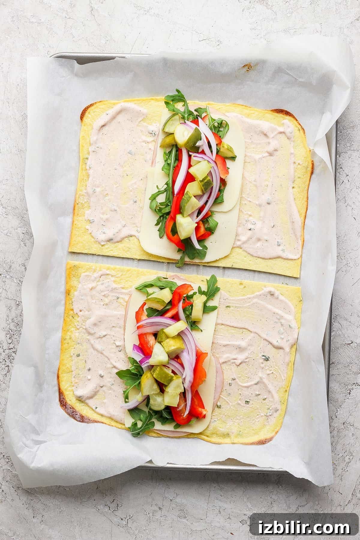 Protein-Packed Cottage Cheese Wraps Recipe 4 Two open-faced flatbread sandwiches on a baking sheet, with cheese, arugula, red onions, red peppers, and pickles, topped with a creamy spread, placed on parchment paper.