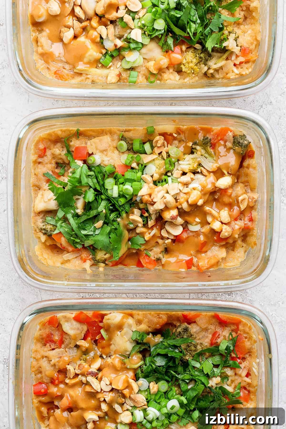Baked Peanut Butter Chicken for Meal Prep 2 Three glass containers filled with a delicious dish featuring rice, diced vegetables, chopped nuts, and tender peanut chicken, all garnished with fresh herbs and green onions.