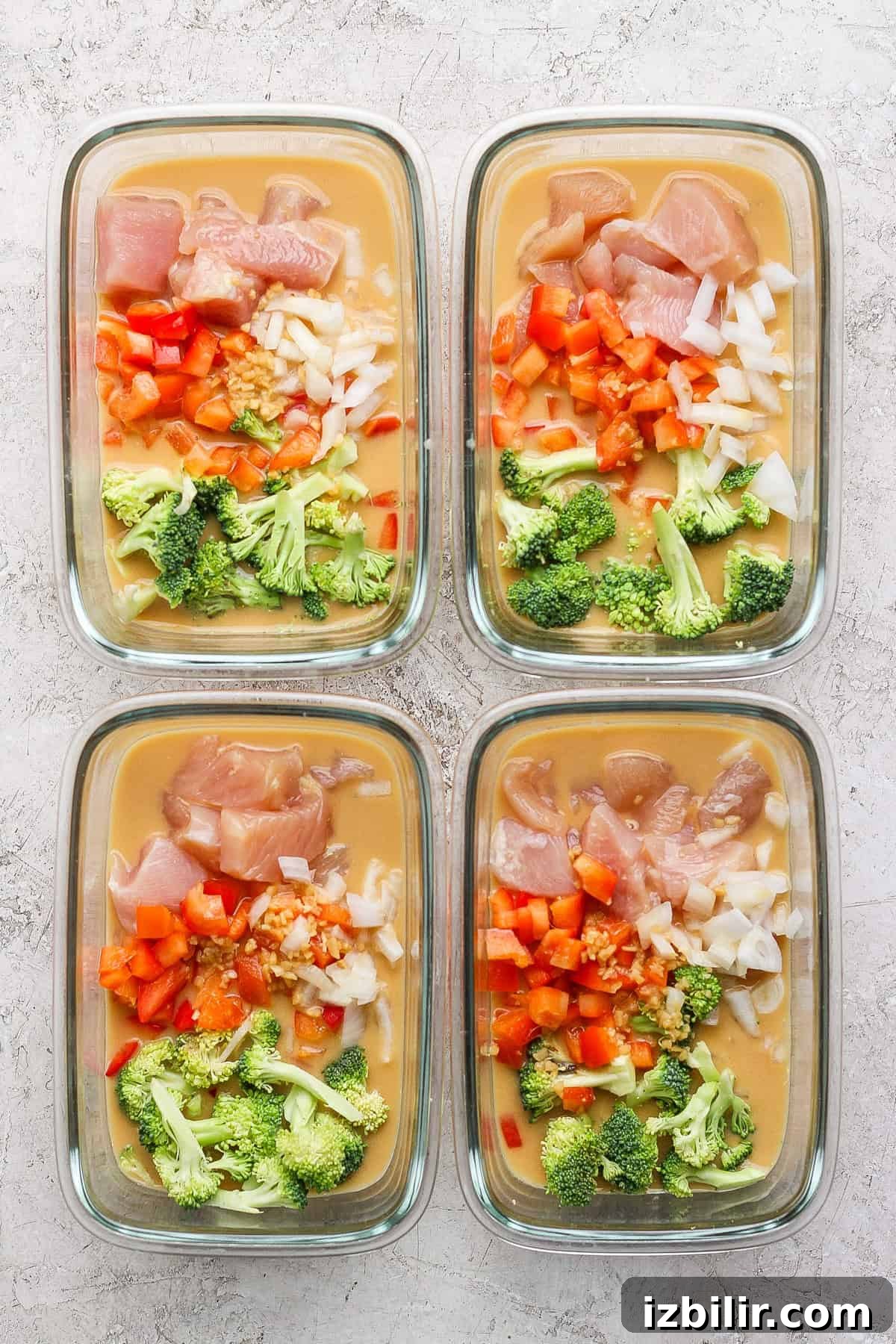 Baked Peanut Butter Chicken for Meal Prep 3 Four glass meal prep containers filled with uncooked peanut chicken, diced peppers, onions, broccoli, and sauce rest on a textured surface.