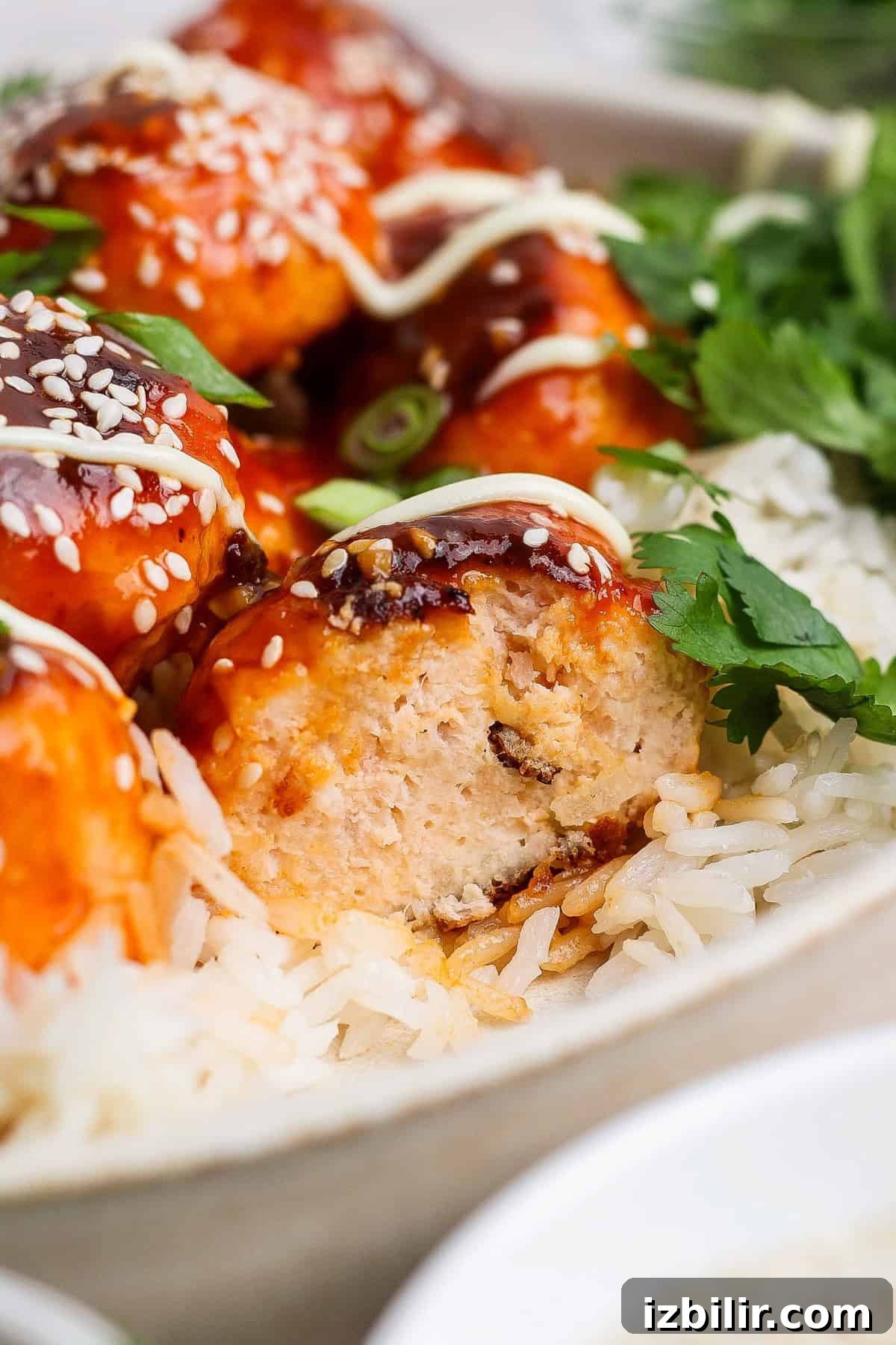 Sesame Ginger Salmon Meatballs with Sticky BBQ Glaze 5 Close-up of rice topped with saucy chicken meatballs, garnished with sesame seeds and cilantro. A meatball is cut open, revealing its interior. Drizzled sauce is visible on top.