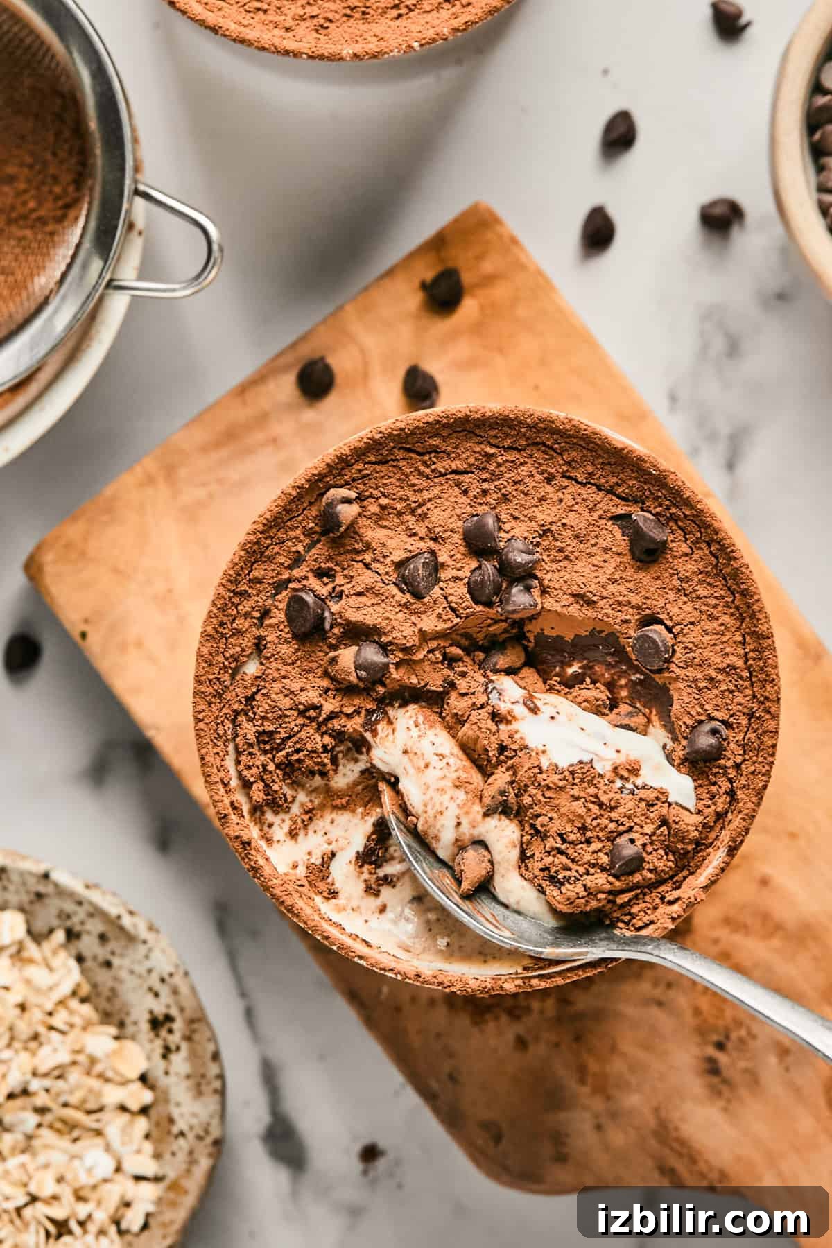 Bowl of tiramisu topped with cocoa powder and chocolate chips on a wooden board, next to a sieve, oats, and scattered chocolate chips. A spoon is scooping a portion from the bowl.