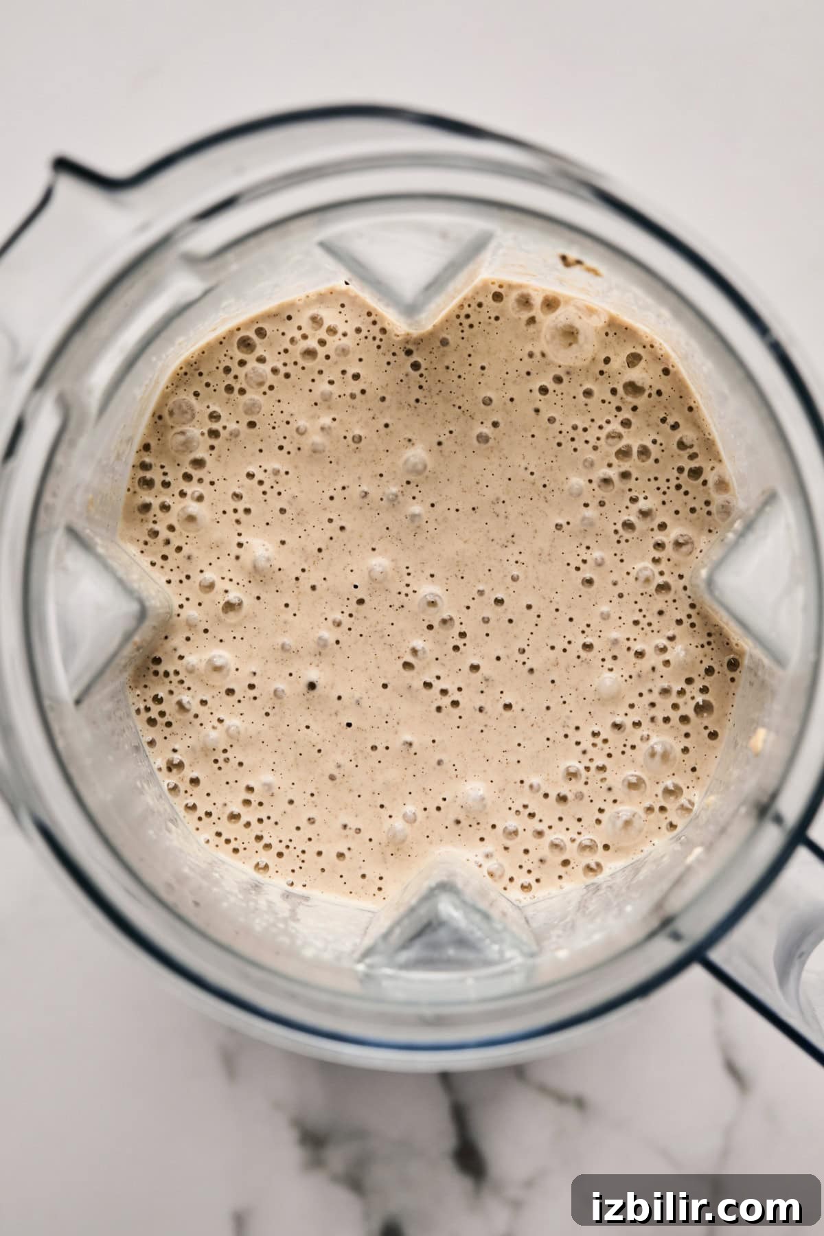 Top view of a blender filled with a frothy beige liquid, possibly a smoothie, on a white marble surface.