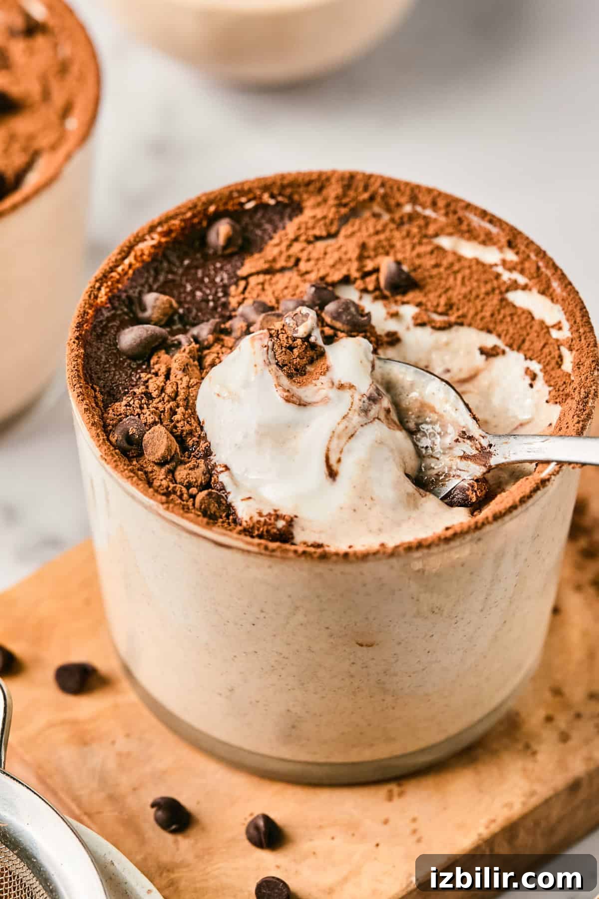 Tiramisu in a ceramic cup topped with cocoa powder and chocolate chips, with a spoon scooping a portion.