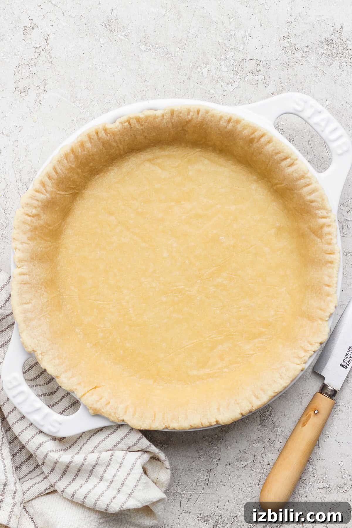 Protein Packed Pie Crust Recipe 2 Unbaked pie crust in a white pie dish on a textured surface with a knife and striped cloth nearby.