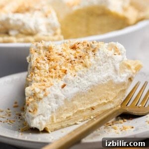 Protein Packed Pie Crust Recipe 3 A slice of coconut cream pie on a white plate with a gold fork, topped with whipped cream and toasted coconut flakes.