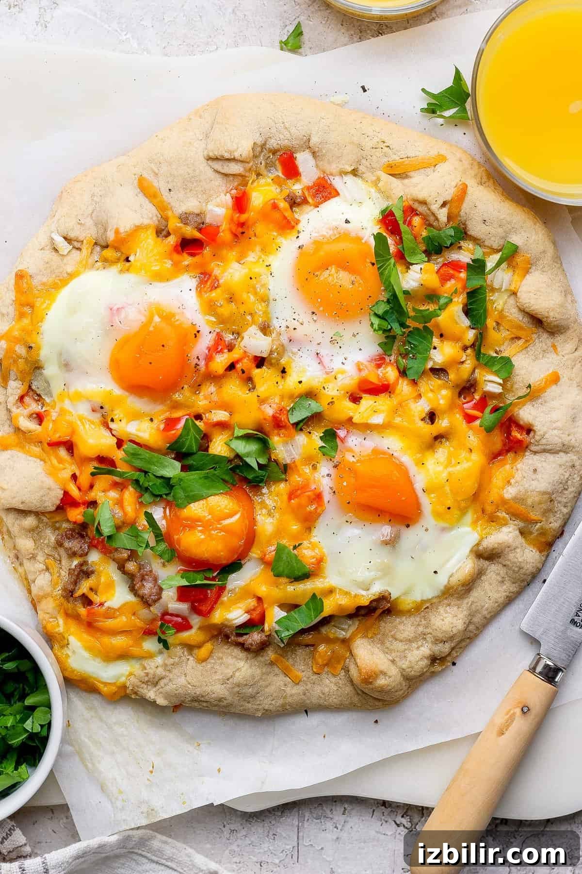 A baked breakfast pizza topped with sunny-side-up eggs, cheese, red peppers, and herbs on parchment paper, with a bowl of chopped parsley and a glass of orange juice nearby.