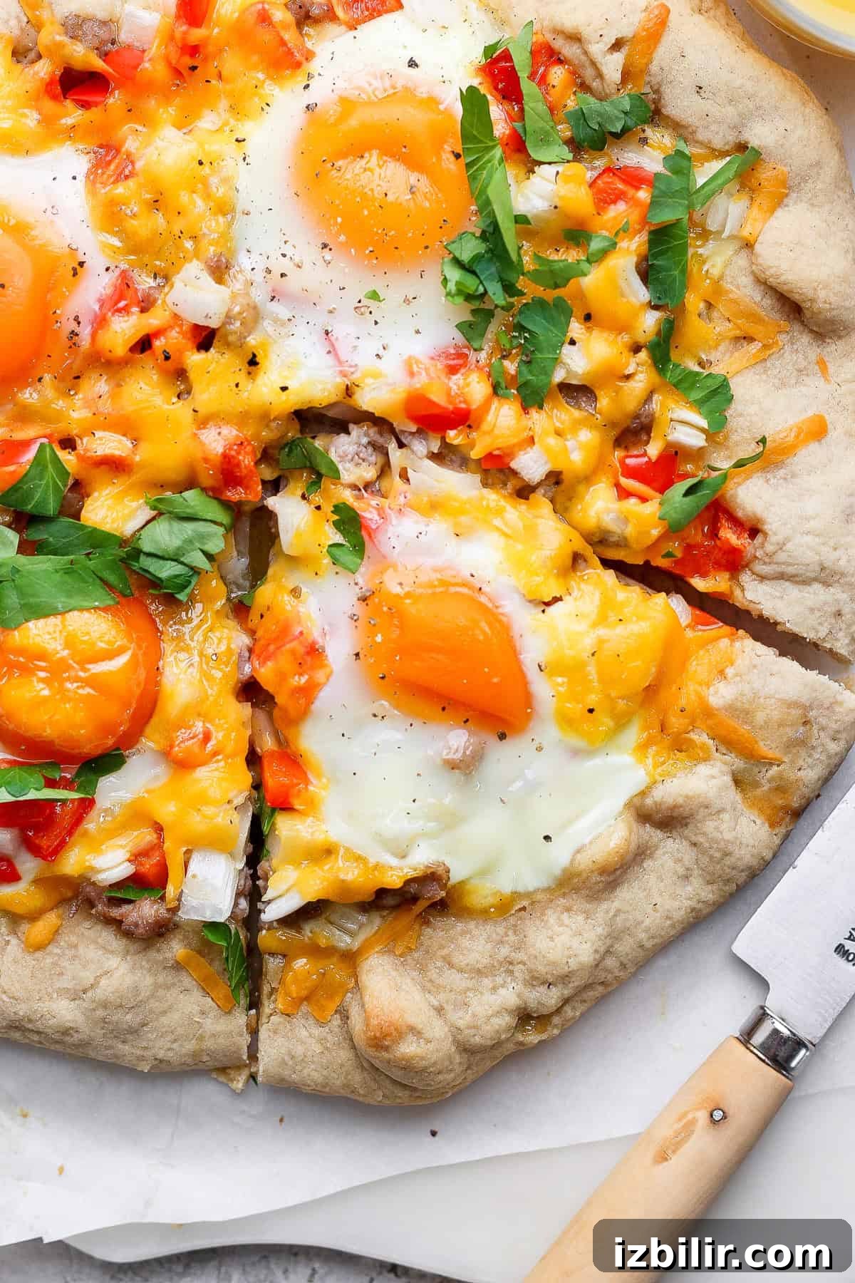 A pizza topped with sunny-side-up eggs, melted cheese, diced red bell peppers, onions, and fresh herbs. A slice is cut out, with a knife resting nearby.