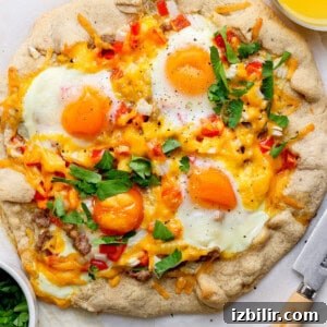 A rustic breakfast pizza topped with sunny-side-up eggs, cheese, diced red peppers, and fresh parsley on a light crust, placed next to a knife and a bowl of chopped greens.