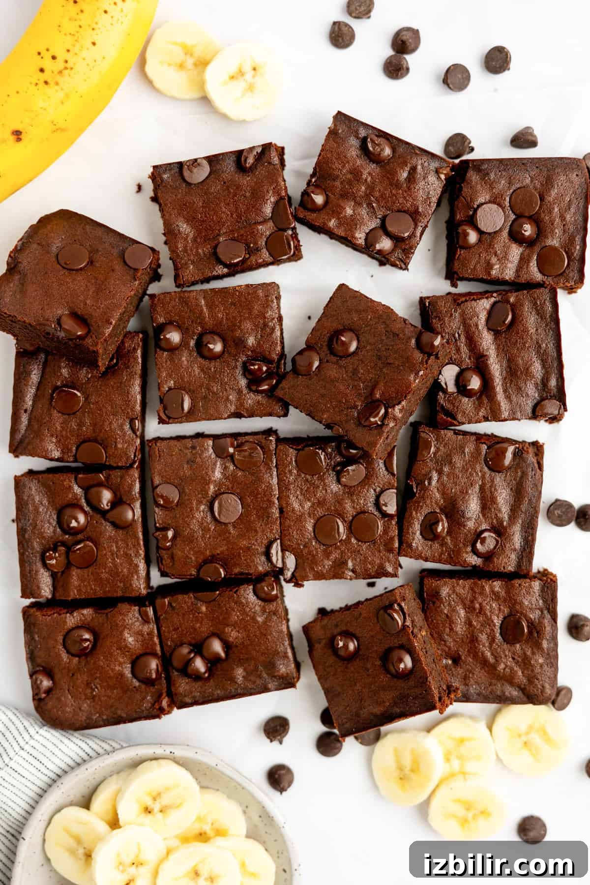 Brownies with chocolate chips arranged on a surface, surrounded by banana slices and a whole banana.