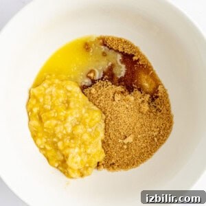 A white bowl containing mashed bananas, brown sugar, and melted butter on a white surface.