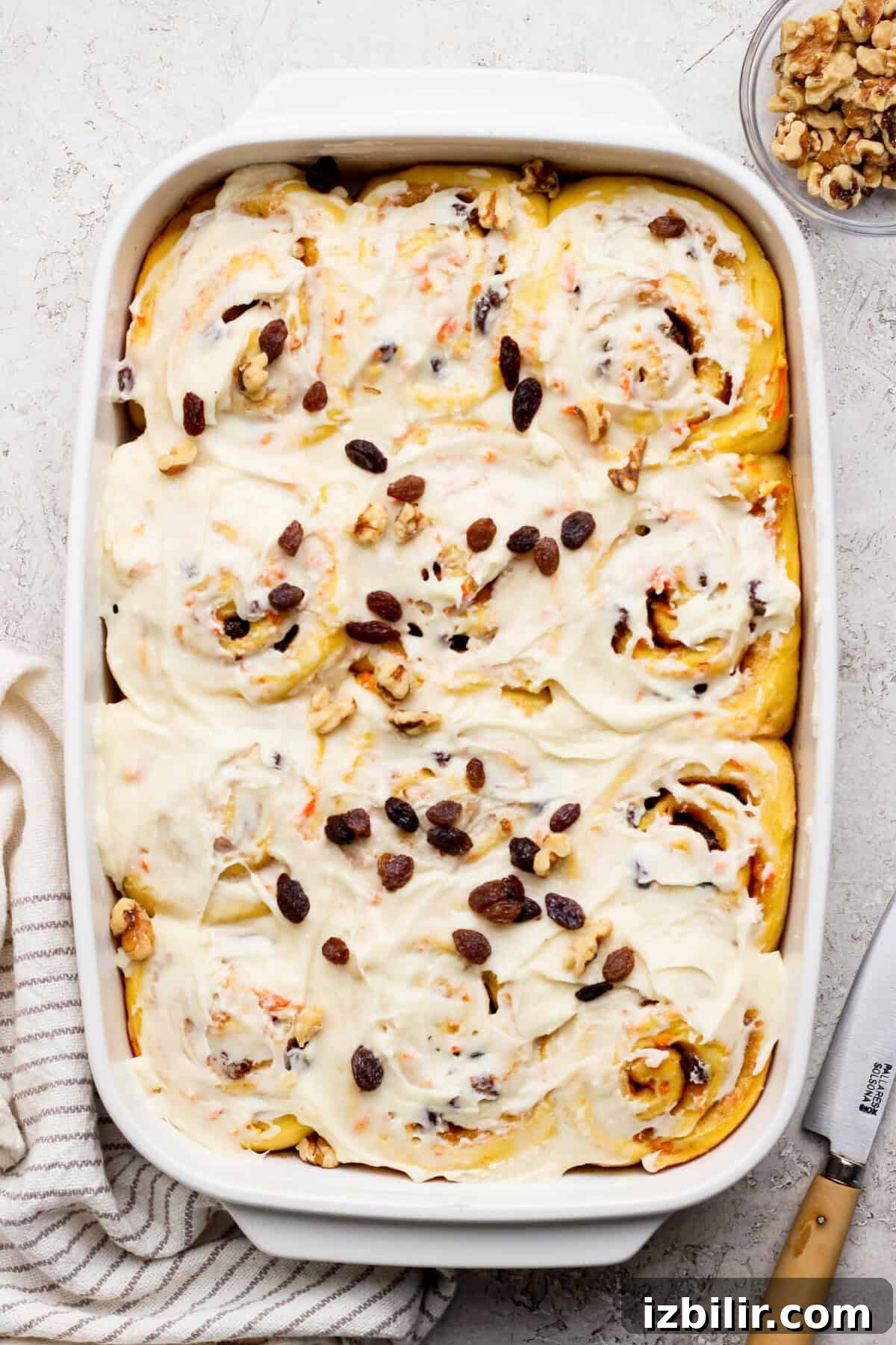 Carrot Cake Cinnamon Rolls Recipe with Cream Cheese Frosting 2 A baking dish filled with frosted carrot cake cinnamon rolls topped with raisins and walnuts. A knife and striped cloth are nearby.