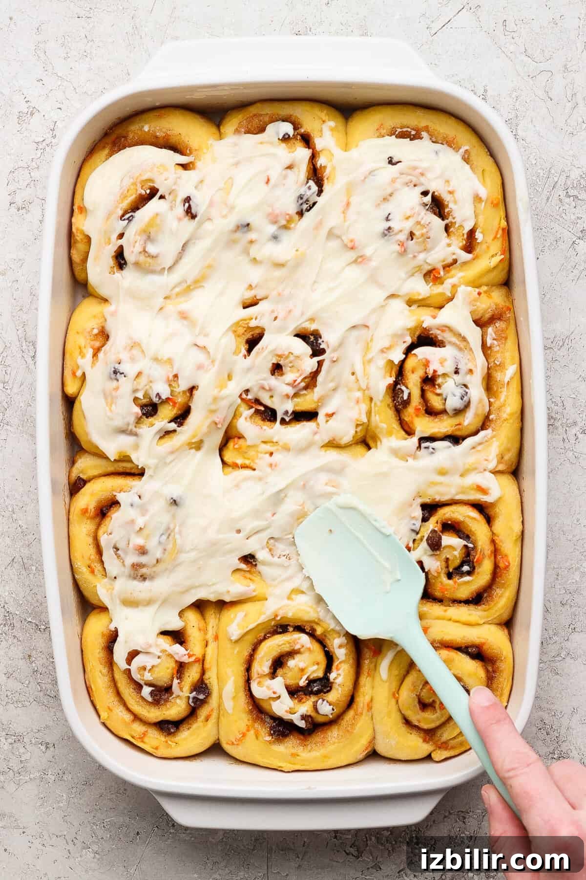 Carrot Cake Cinnamon Rolls Recipe with Cream Cheese Frosting 5 A person spreads white icing over freshly baked carrot cake cinnamon rolls arranged in a rectangular baking dish.