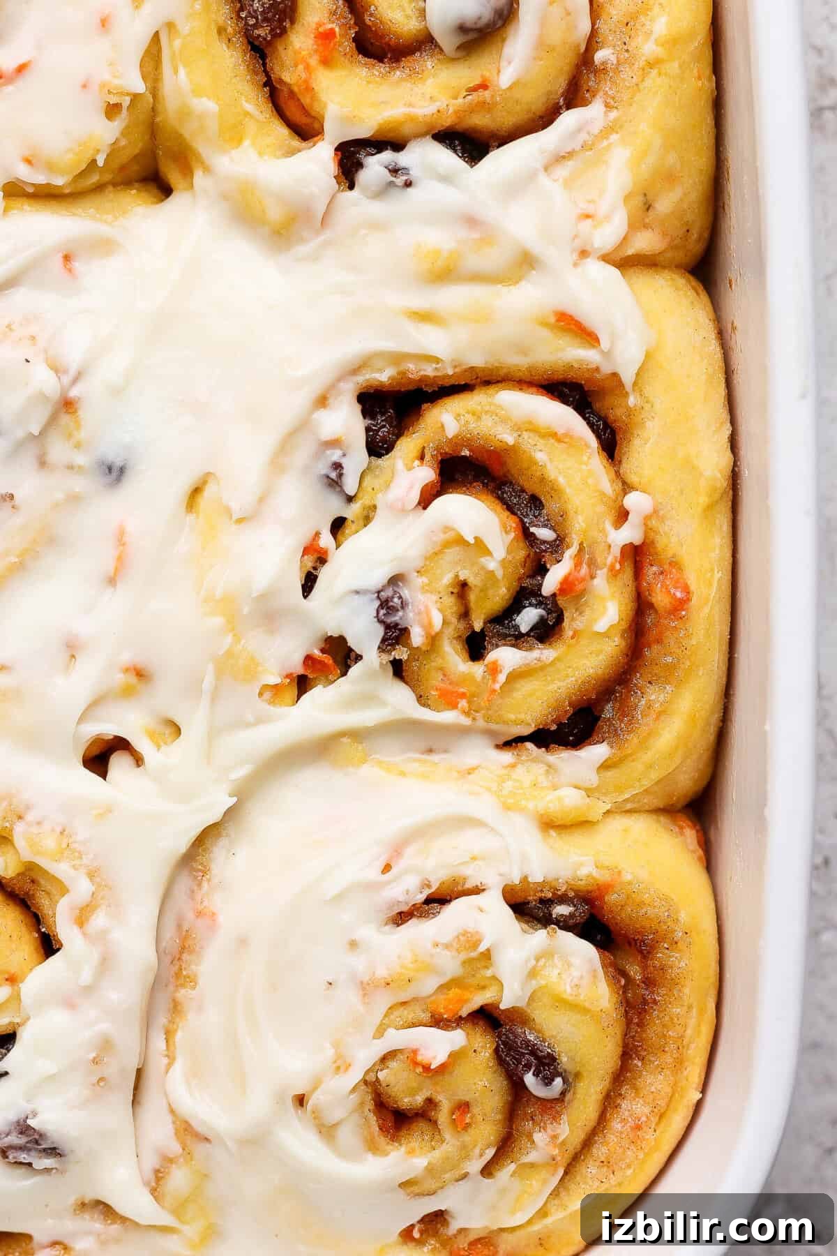 Carrot Cake Cinnamon Rolls Recipe with Cream Cheese Frosting 6 Close-up of frosted carrot cake cinnamon rolls in a baking dish, featuring swirls of dough with cinnamon and raisin filling, topped with luscious cream cheese icing.