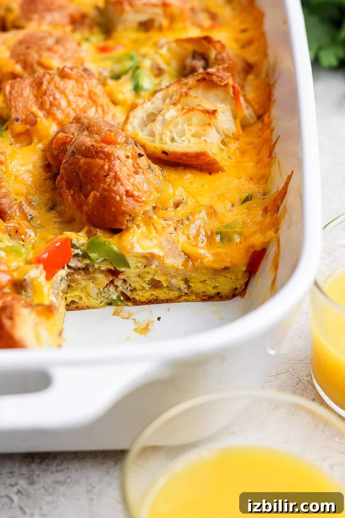 A baked breakfast casserole in a white dish, featuring melted cheese, vegetables, and biscuit pieces with two glasses of orange juice beside it.
