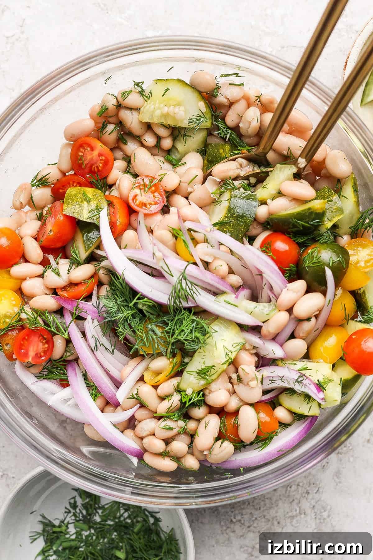 A bowl of bean salad with white beans, cherry tomatoes, red onion, cucumber, fresh dill, and dressing, garnished with herbs, with serving utensils.