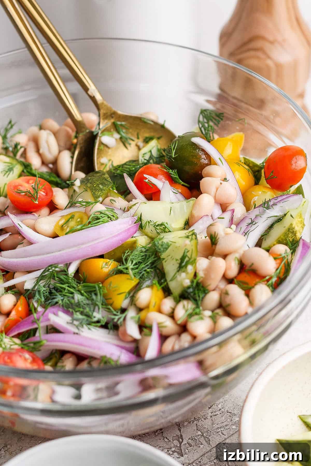 A bowl of vegetable salad with cherry tomatoes, cucumber, white beans, red onion, and fresh dill, tossed with two gold utensils.