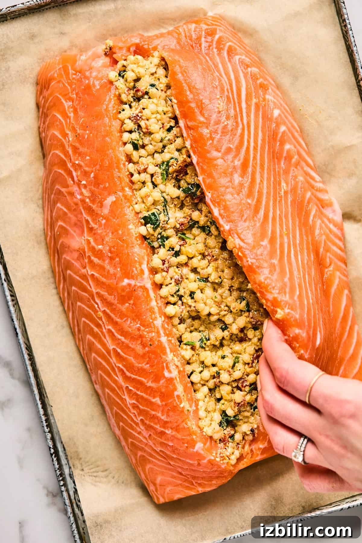 Raw salmon fillet is slit open and filled with a mixture of herbs and grains on a parchment-lined baking tray. A hand adjusts the fillet.