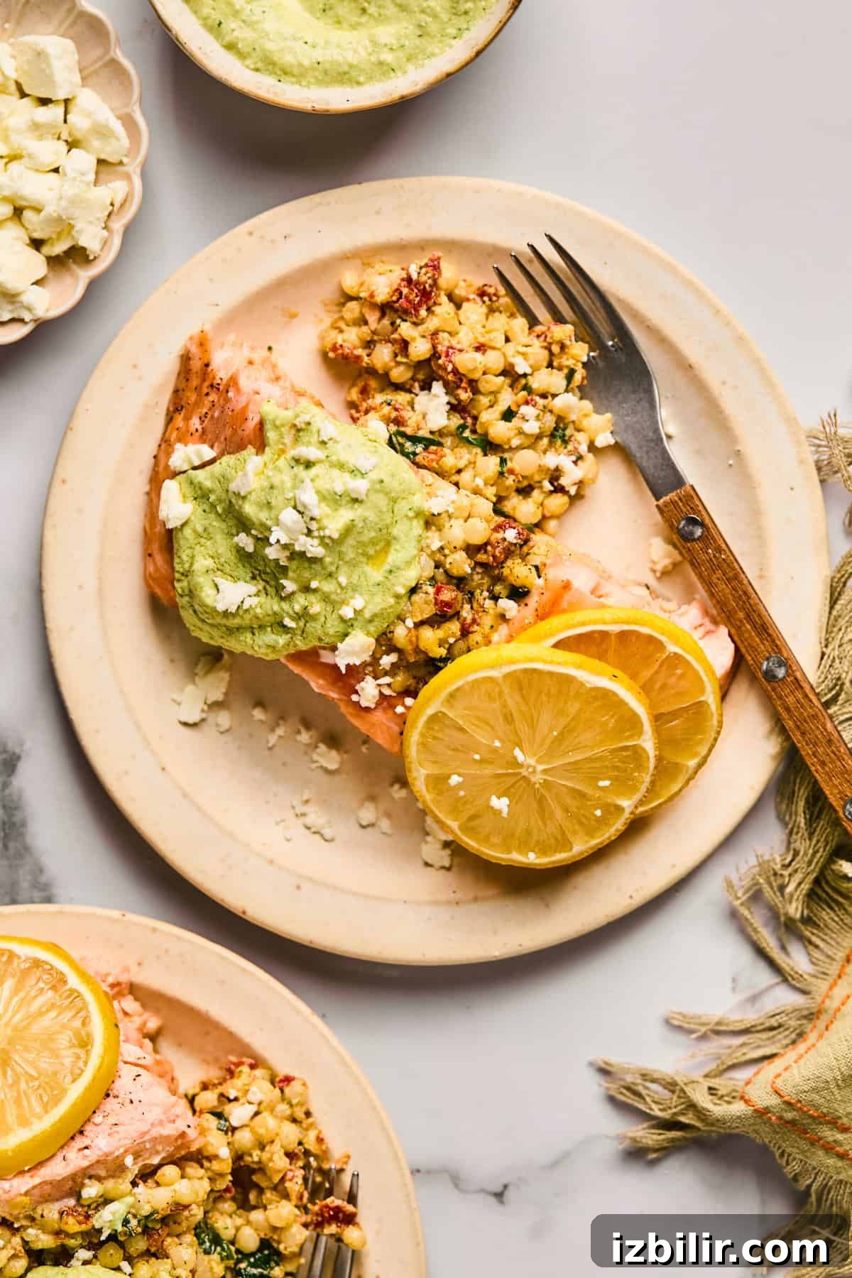 Plate with salmon topped with green sauce, alongside couscous, crumbled cheese, and lemon slices. Fork on the plate, napkin beside it.