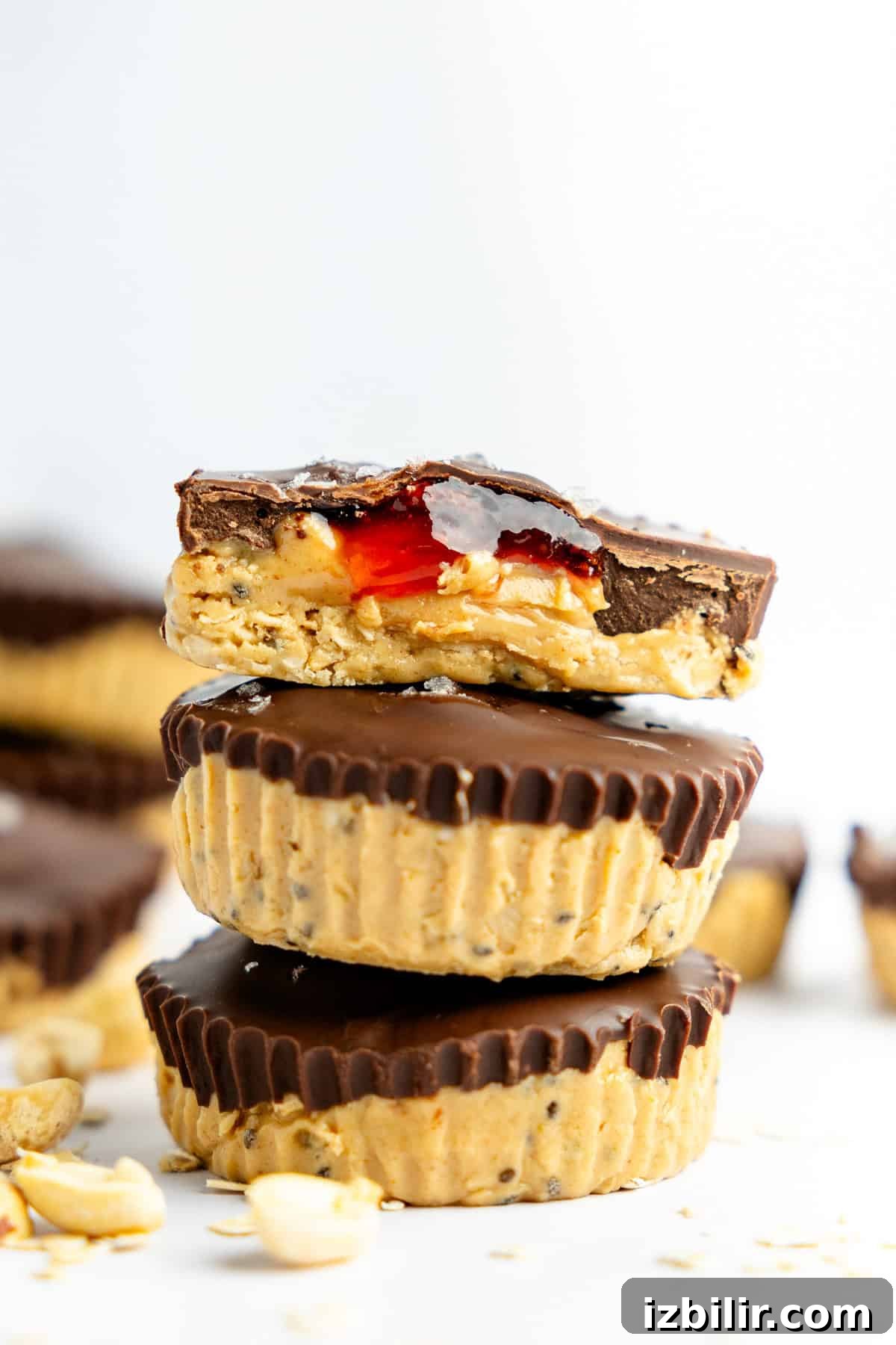 Peanut Butter and Jelly Protein Oatmeal Cups 2 Three peanut butter and chocolate cups stacked, with a bite taken from the top one, revealing a red filling. Peanuts scattered around.