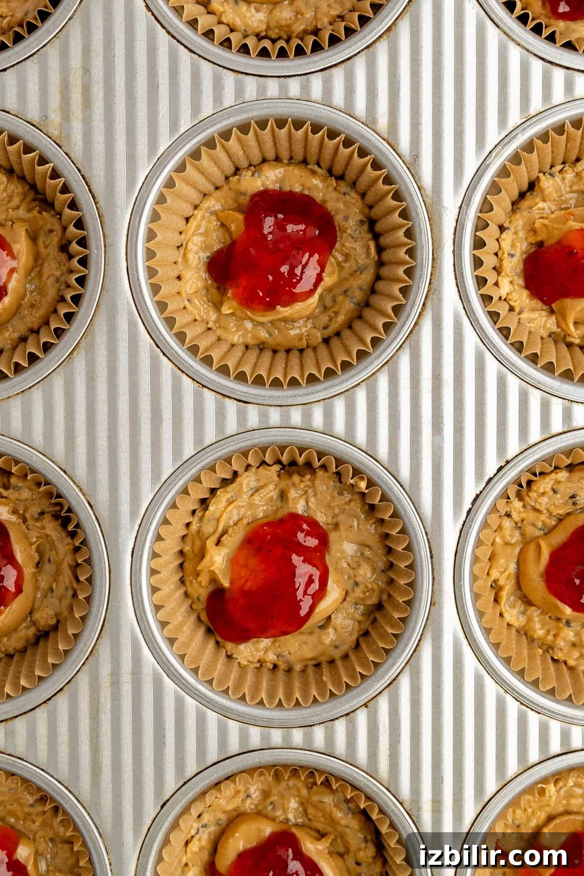 Peanut Butter and Jelly Protein Oatmeal Cups 3 Muffin tin filled with unbaked muffin batter topped with red jam, placed in paper liners, ready for baking.