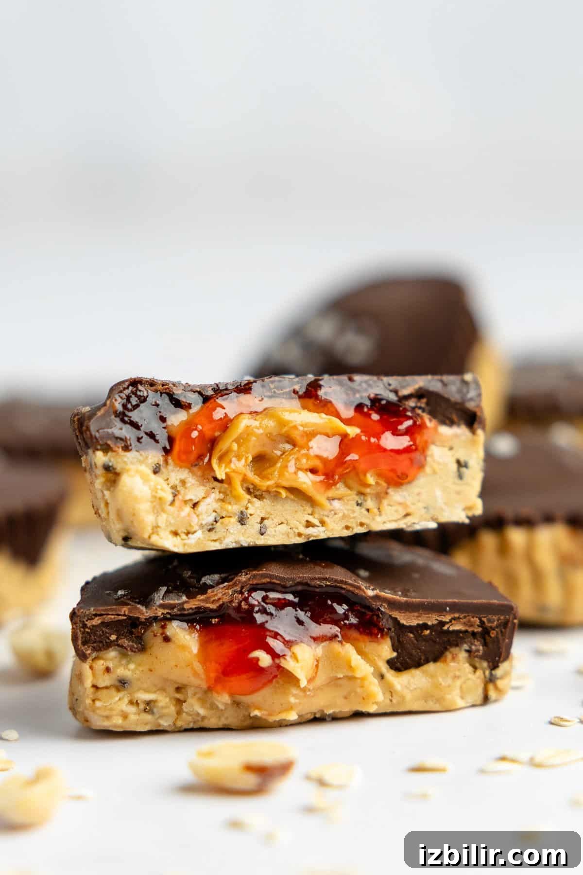 Peanut Butter and Jelly Protein Oatmeal Cups 6 Two chocolate peanut butter cups, one cut in half, revealing peanut butter and jelly filling. Surrounded by more peanut butter cups and scattered oats on a white surface.