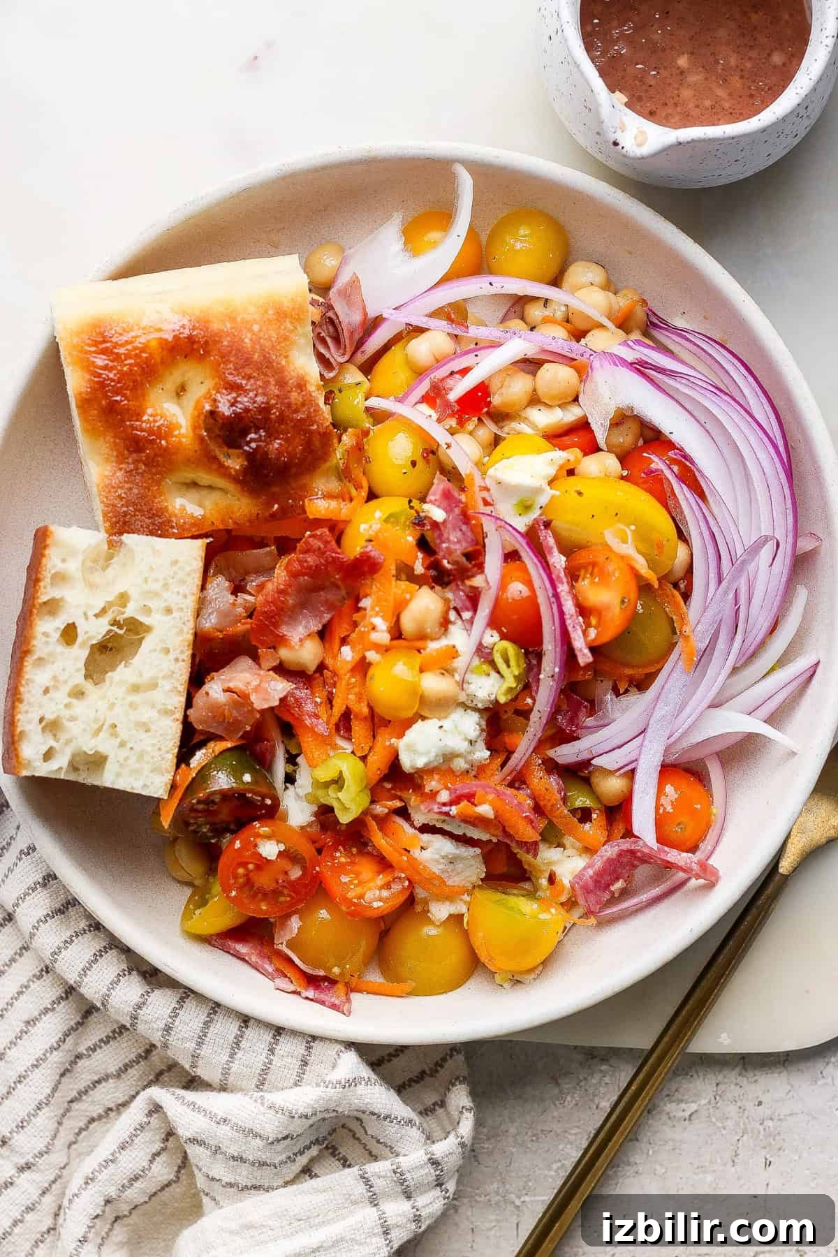 A salad with cherry tomatoes, red onion, carrot shreds, bacon, cheese, and chickpeas, accompanied by two bread pieces. Dressing is in a small cup nearby.